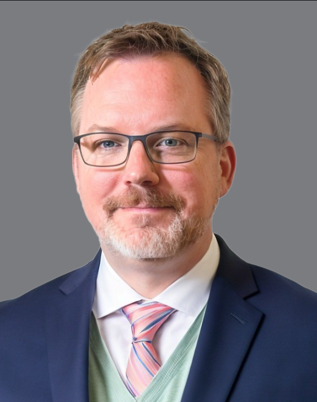 Man with glasses, wearing a blue suit, light green sweater, and pink striped tie, against a gray background.